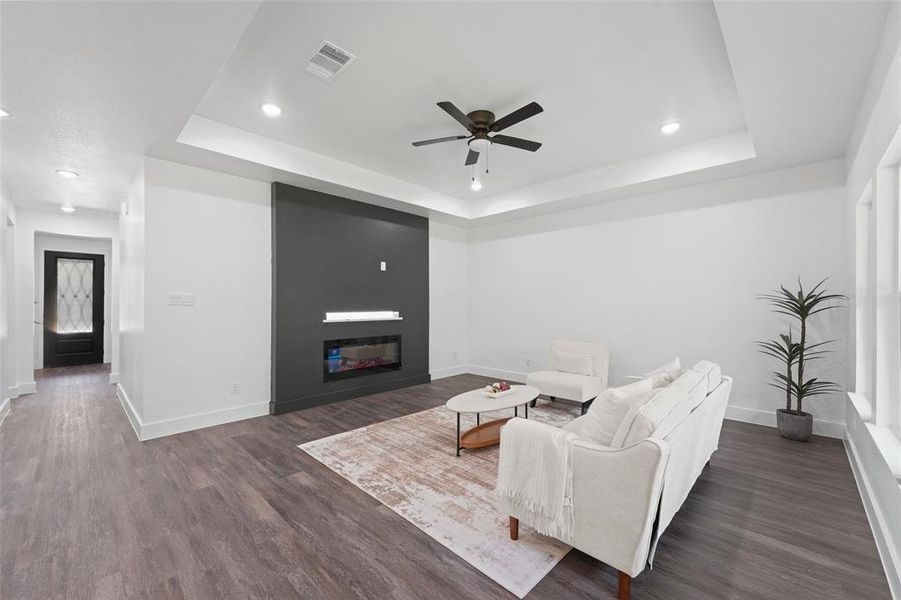 Living area featuring wood-finish flooring, a tray ceiling with a ceiling fan, recessed lighting, and a modern linear fireplace set into a dark accent wall Living area featuring wood-finish flooring, a tray ceiling with a ceiling fan, recessed lighting, and a modern linear fireplace set into a dark accent wall