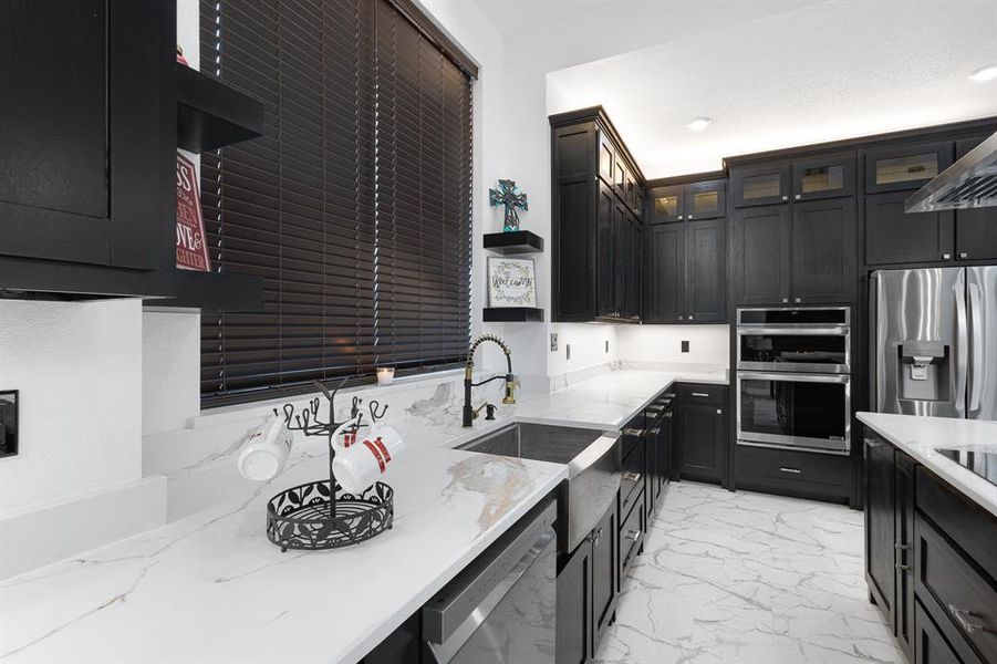 Kitchen featuring dark cabinets, appliances with stainless steel finishes, light stone counters, glass insert cabinets, and light marble finish flooring