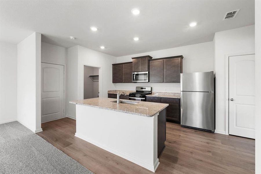 Kitchen featuring dark brown cabinets, appliances with stainless steel finishes, an island with sink, light stone countertops, and recessed lighting