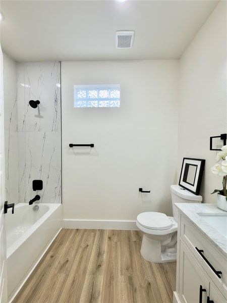 Full bathroom featuring a white bathtub with a tile surround, glass block window, wood-finish flooring, and a white vanity with a countertop