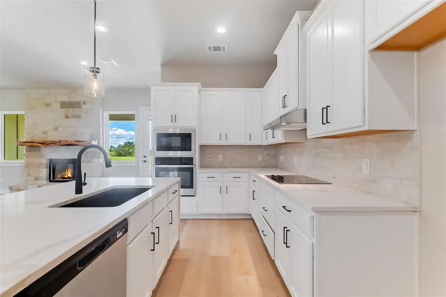 Kitchen featuring decorative light fixtures, light wood-type flooring, stainless steel appliances, a stone fireplace, and white cabinetry
