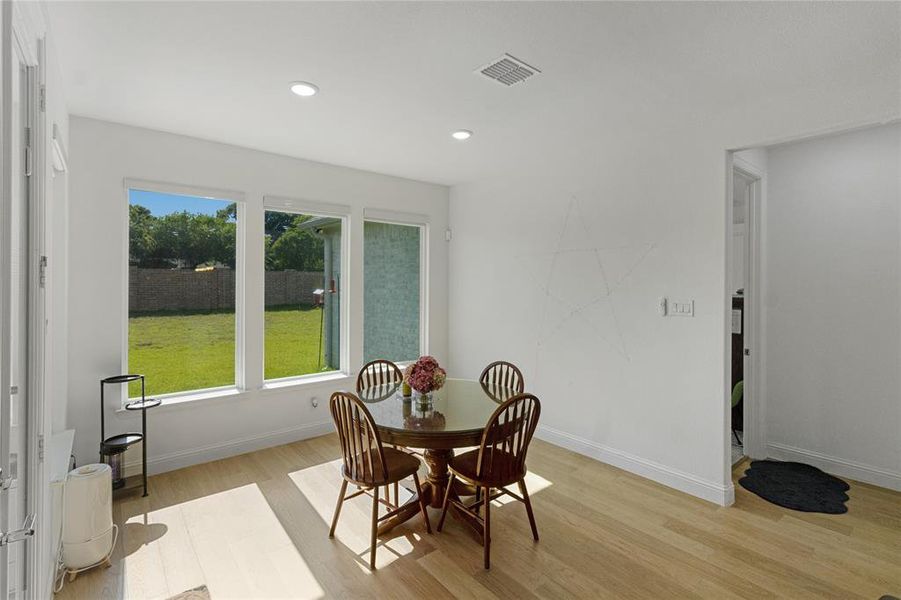 Bright dining area featuring light wood-finish flooring, recessed lighting, and expansive windows showcasing a green lawn and mature perimeter trees