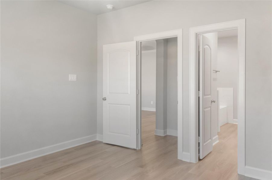 This photo shows a bright, modern room with light wood flooring and neutral-colored walls. It features two white doors; one leads to a bathroom with a visible bathtub, and the other opens to an adjoining room. The space is well-lit and has a clean, minimalistic design.