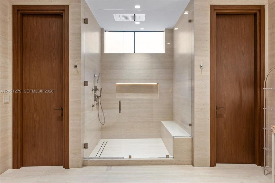 Oversized frameless glass walk-in shower with travertine tile surround, built-in bench, illuminated recessed niche, ceiling rain head, multi-function shower fixtures, clerestory window, and dual walnut entry doors flanking each side.