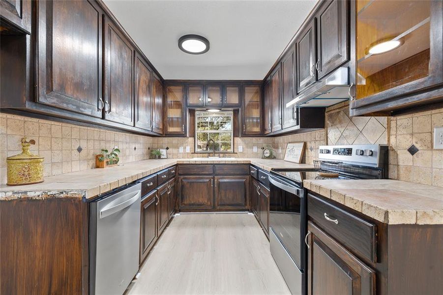 Kitchen with dark brown cabinetry, stainless steel appliances, glass insert cabinets, decorative backsplash, and under cabinet range hood