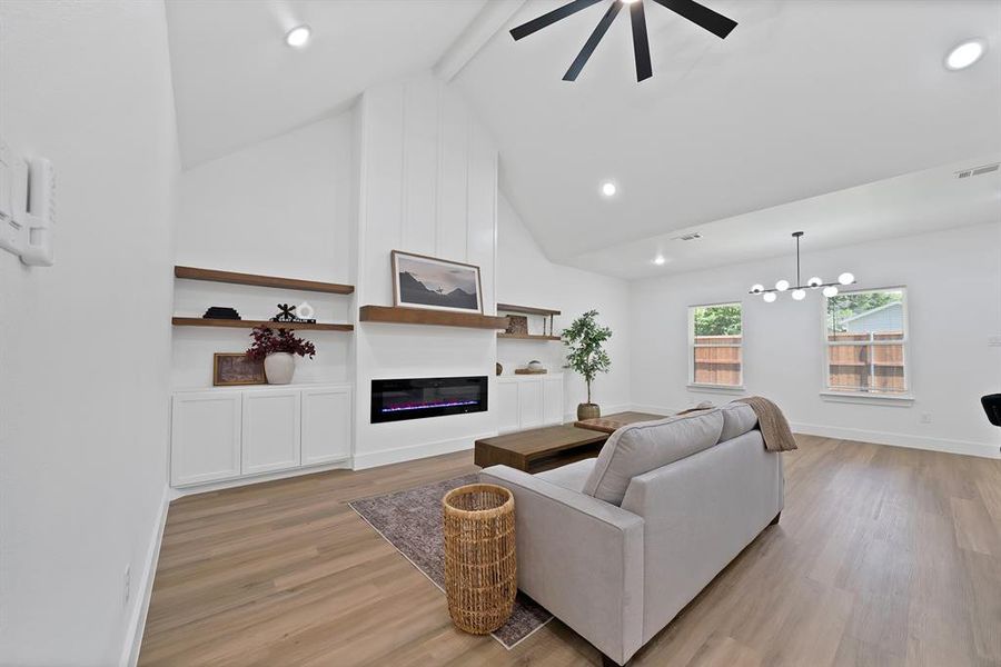Spacious living area featuring a vaulted ceiling with recessed lighting and a ceiling fan, a contemporary linear electric fireplace, wood-finish flooring, and built-in shelving with lower cabinetry Spacious living area featuring a vaulted ceiling with recessed lighting and a ceiling fan, a contemporary linear electric fireplace, wood-finish flooring, and built-in shelving with lower cabinetry