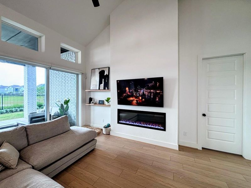 Bright and modern living room with high ceilings, featuring a sleek electric fireplace and mounted TV. Large windows and sliding glass doors offer abundant natural light and views of the backyard.