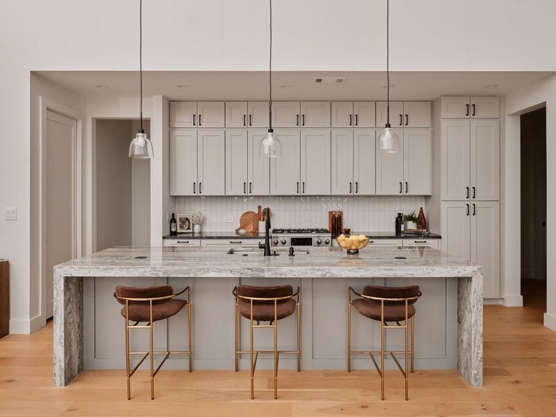 Modern kitchen featuring light wood-finish flooring, a waterfall island with a patterned countertop, light gray cabinetry, a subway tile backsplash, and three clear glass pendant lights