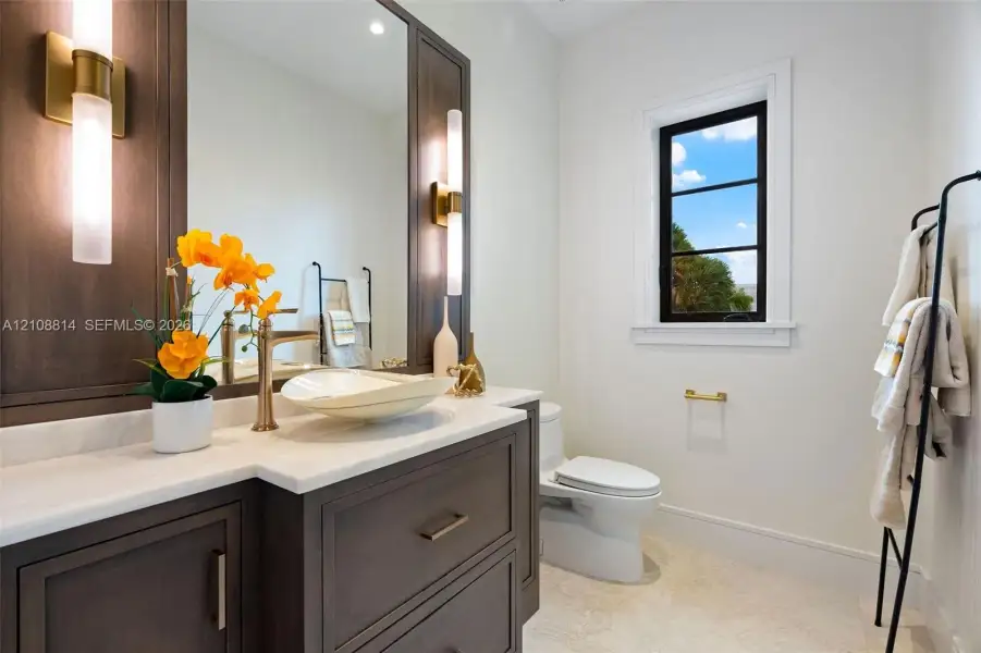 Dark wood vanity featuring a light-toned stone countertop, vessel sink, brass-finish hardware, integrated vertical sconces, and a multi-pane window Dark wood vanity featuring a light-toned stone countertop, vessel sink, brass-finish hardware, integrated vertical sconces, and a multi-pane window