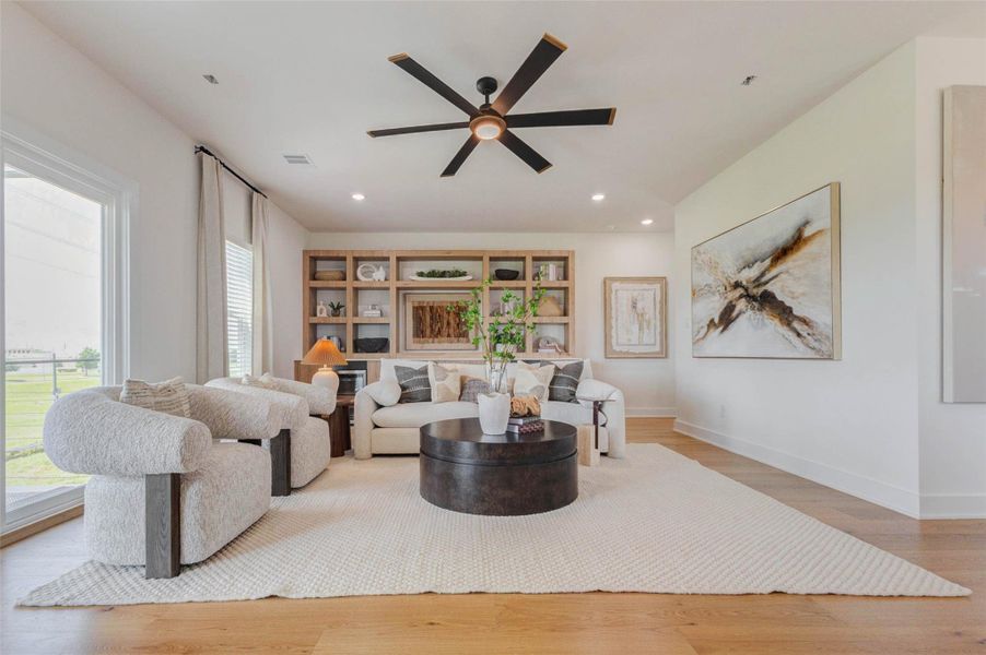 A beautifully proportioned living room offers an inviting setting for relaxation and entertaining. Curved upholstered seating, a generous sofa, and a bold round coffee table create a comfortable conversation area beneath a modern ceiling fan. Custom wood shelving provides display and storage space, while oversized artwork adds visual impact. The neutral palette, warm flooring, and soft textures create a sophisticated yet welcoming atmosphere.