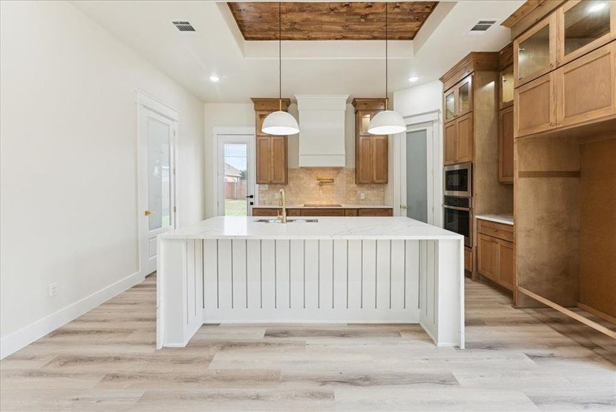Kitchen featuring a raised ceiling, brown cabinets, light wood-type flooring, light stone counters, and recessed lighting