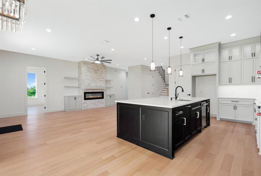 Spacious kitchen and living area featuring wood-finish flooring, a central kitchen island with a sink and pendant lighting, white shaker cabinetry with tile backsplash, a stacked stone fireplace with built-in shelving and cabinetry, and recessed lighting throughout Spacious kitchen and living area featuring wood-finish flooring, a central kitchen island with a sink and pendant lighting, white shaker cabinetry with tile backsplash, a stacked stone fireplace with built-in shelving and cabinetry, and recessed lighting throughout