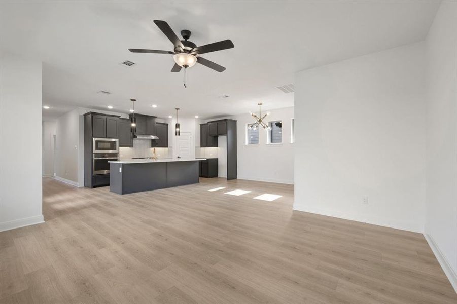 Open-concept interior featuring wood-finish flooring, a modern kitchen with dark cabinetry, an island with countertop seating, built-in stainless steel appliances, and contemporary pendant lighting