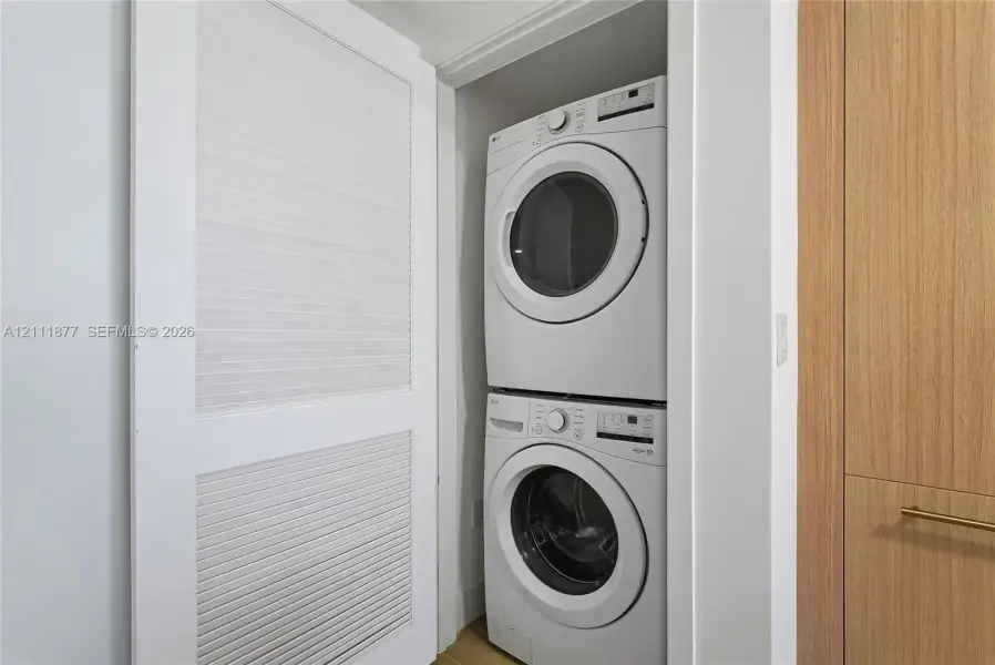 Stacked laundry unit featuring white appliances, enclosed by white louvered bifold doors and wood-finish cabinetry