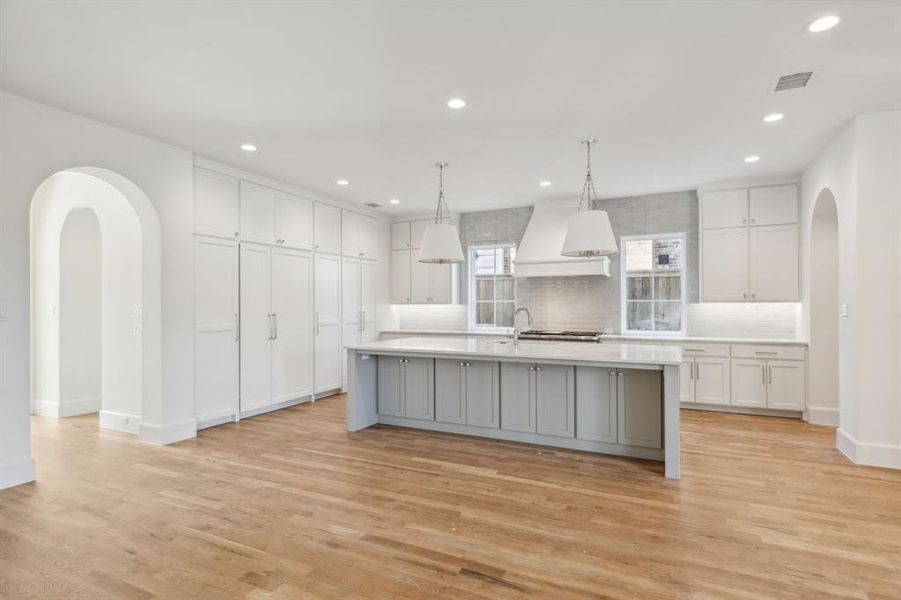 Kitchen featuring backsplash, hanging light fixtures, light hardwood flooring, premium range hood, and a center island with sink