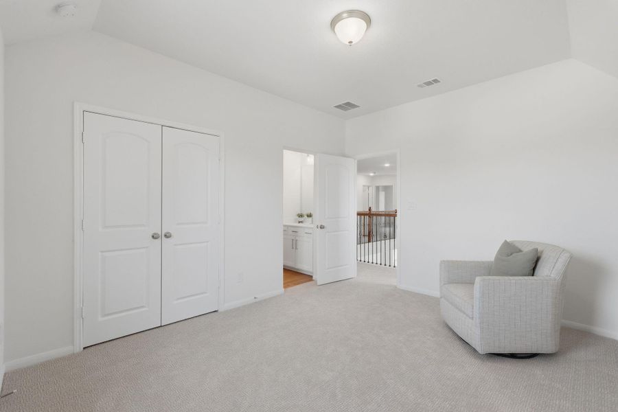 This photo shows a bright, carpeted room with neutral walls, featuring a double closet and a cozy armchair. An open doorway leads to a hallway with a railing, suggesting an upper-level space in the home.