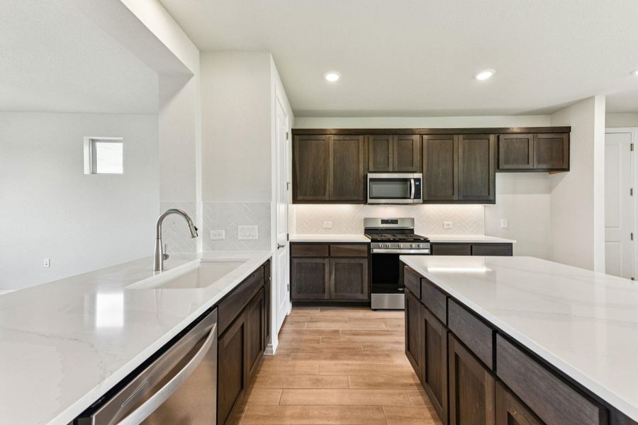 Kitchen with dark brown cabinets, decorative backsplash, stainless steel appliances, light stone counters, and light wood-style floors