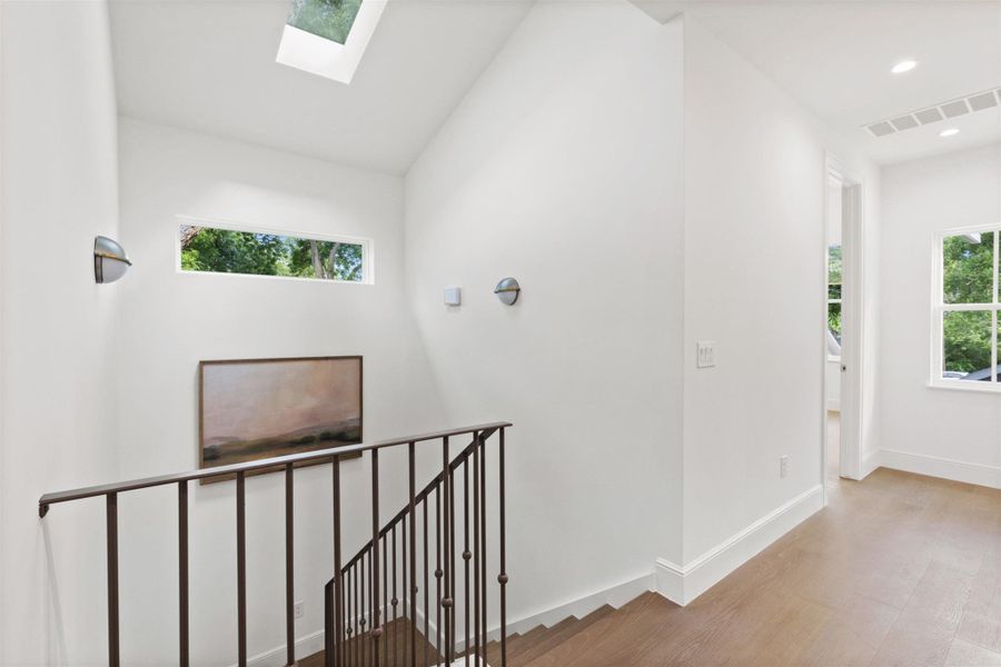 A luminous upper landing features rich hardwood floors and an open staircase, illuminated by a skylight and a high horizontal window.