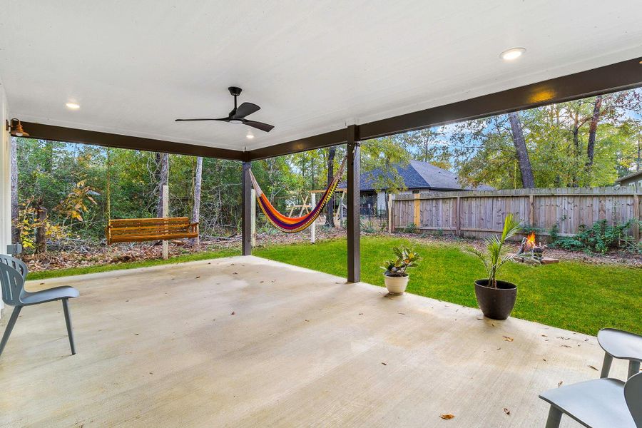 This photo showcases a spacious, covered patio with a ceiling fan, perfect for relaxing outdoors. It features a hammock and seating area, overlooking a lush backyard with a wooden swing, all surrounded by a privacy fence. Ideal for entertaining and enjoying nature.
