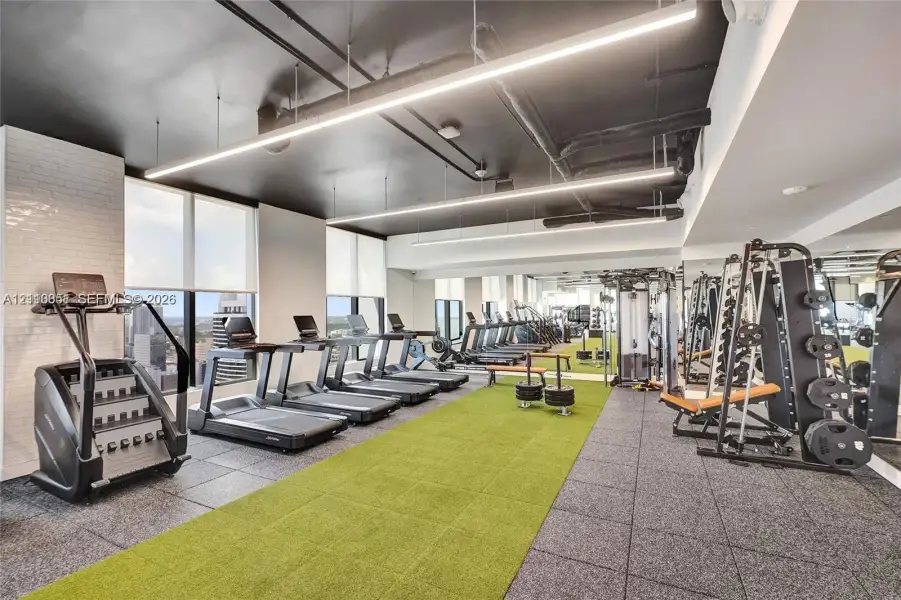 Fitness facility featuring floor-to-ceiling windows, high-contrast exposed industrial-style ceiling, integrated linear LED lighting, wall-to-wall mirrored surfaces, and a combination of rubber and artificial turf flooring