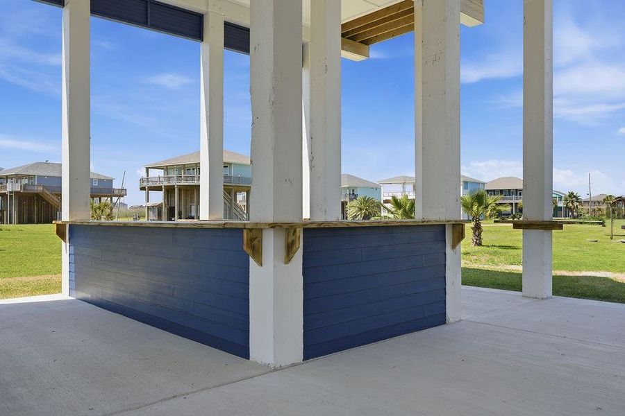 This photo shows an outdoor covered area with a bar-style counter, surrounded by open green space and neighboring elevated homes, ideal for entertaining and enjoying coastal living.