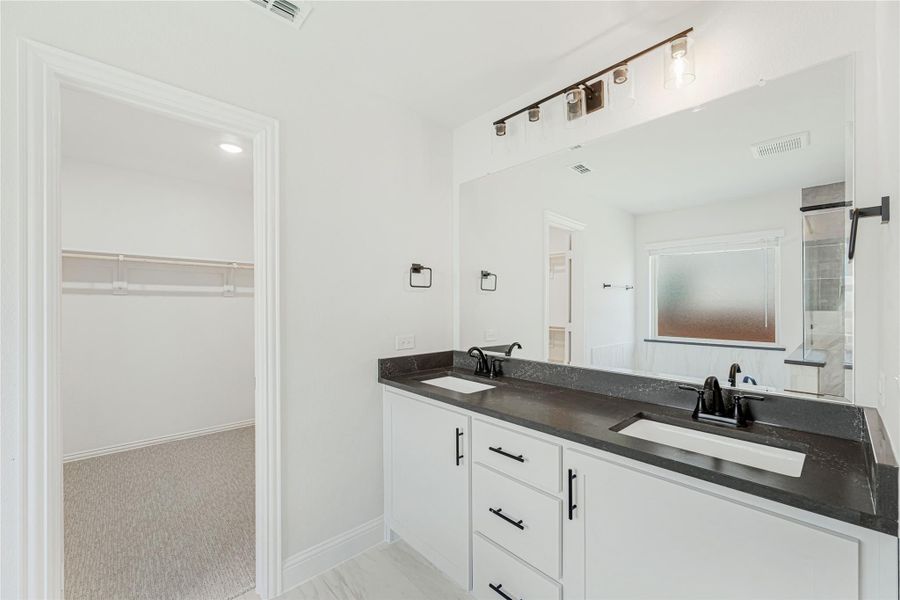 Bathroom with double sink vanity, dark countertops, white cabinets, and walk-in closet entrance