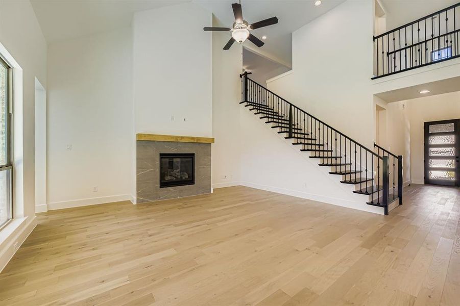 Unfurnished living room with high vaulted ceiling, light wood-style floors, a tiled fireplace, stairs, and recessed lighting Unfurnished living room with high vaulted ceiling, light wood-style floors, a tiled fireplace, stairs, and recessed lighting