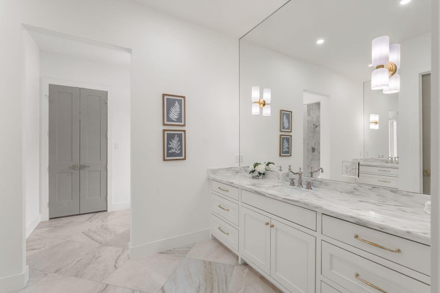 Stunning primary bath with an oversized dual-sink vanity topped in marble and framed by a full wall mirror that reflects natural light throughout the space. Brass hardware and designer sconces add warmth against crisp white cabinetry.