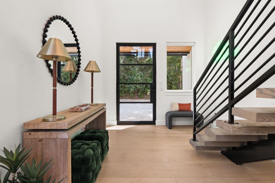 This entryway features modern design elements with a black metal staircase, light wood flooring, and a large windowed door. Natural light and greenery from outside enhance the inviting atmosphere.