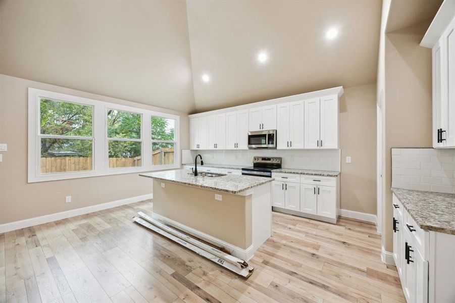 Kitchen with backsplash, a kitchen island with sink, white cabinets, light stone counters, and high vaulted ceiling Kitchen with backsplash, a kitchen island with sink, white cabinets, light stone counters, and high vaulted ceiling