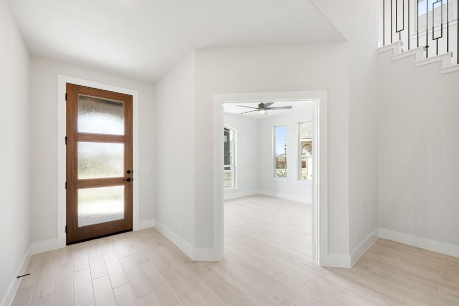 Bright entryway with wood and glass front door, light hardwood floors, and open view to adjacent room with ceiling fan.