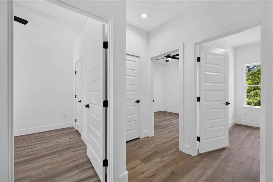 Hallway featuring wood-finish flooring, white paneled doors with black hardware, and crisp white walls Hallway featuring wood-finish flooring, white paneled doors with black hardware, and crisp white walls