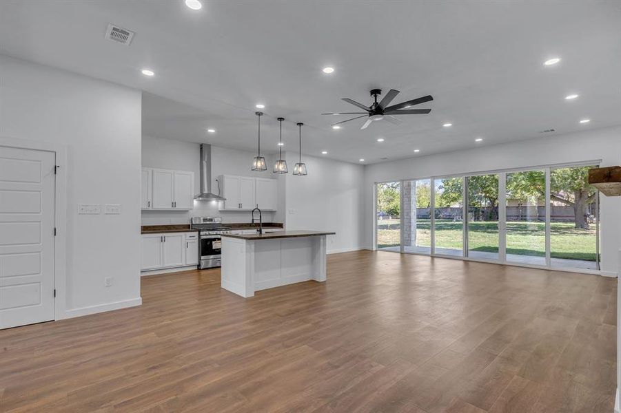 Kitchen featuring a center island with sink, wall chimney range hood, hanging light fixtures, stainless steel range, and white cabinets