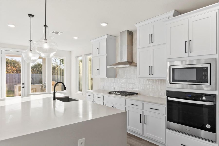 Bright, modern kitchen with white cabinetry and sleek black hardware. Features a large island with a dark faucet and pendant lighting. Includes built-in stainless steel appliances and a cooktop. Ample natural light from large windows and glass doors leading to the backyard. Bright, modern kitchen with white cabinetry and sleek black hardware. Features a large island with a dark faucet and pendant lighting. Includes built-in stainless steel appliances and a cooktop. Ample natural light from large windows and glass doors leading to the backyard.