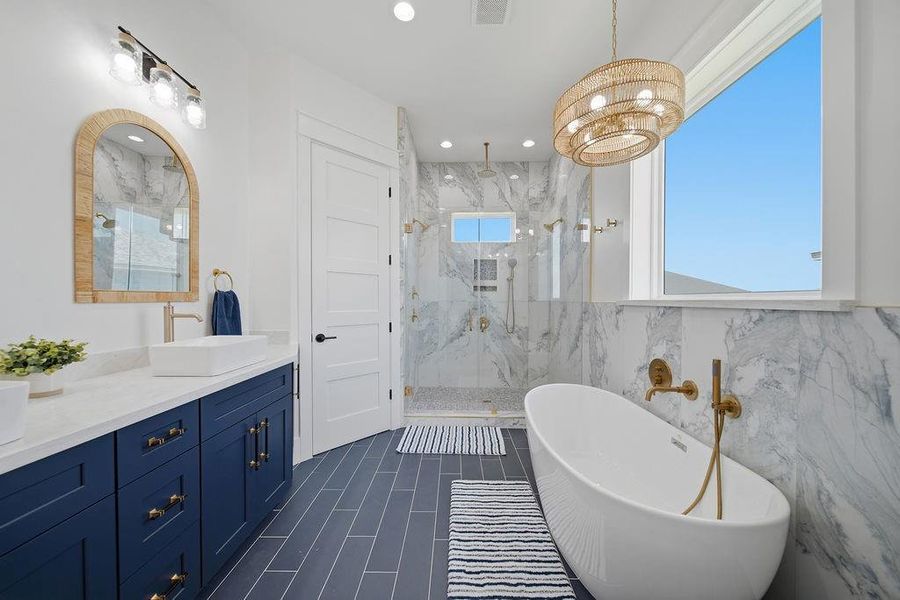 This bathroom features a modern design with a freestanding tub, glass-enclosed shower, and elegant marble tiles. It includes a double vanity with navy cabinets and gold fixtures, complemented by a large window and stylish lighting.