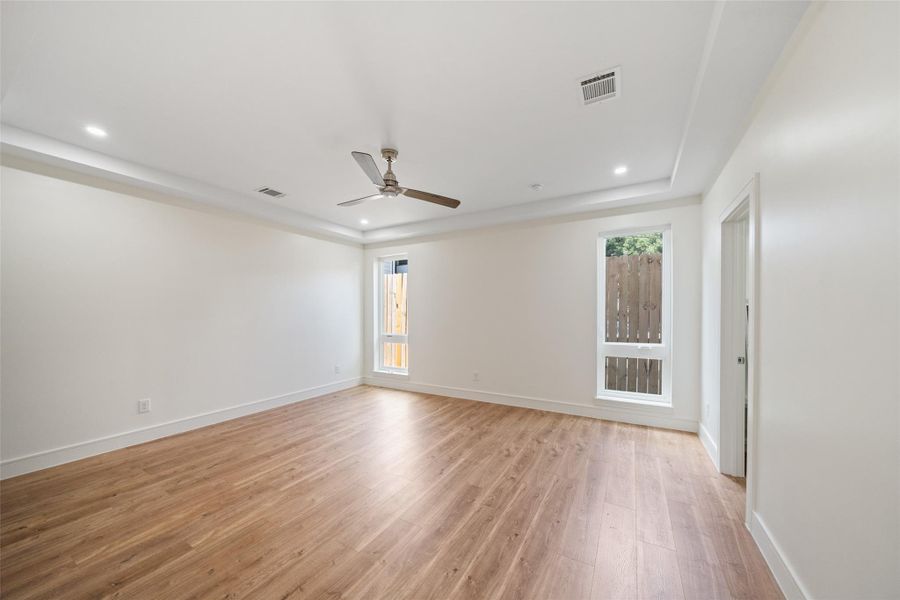 First floor bedroom features clean, white walls and wood flooring, with recessed lighting and a ceiling fan for comfort. Large windows allow natural light, creating a bright and inviting atmosphere.