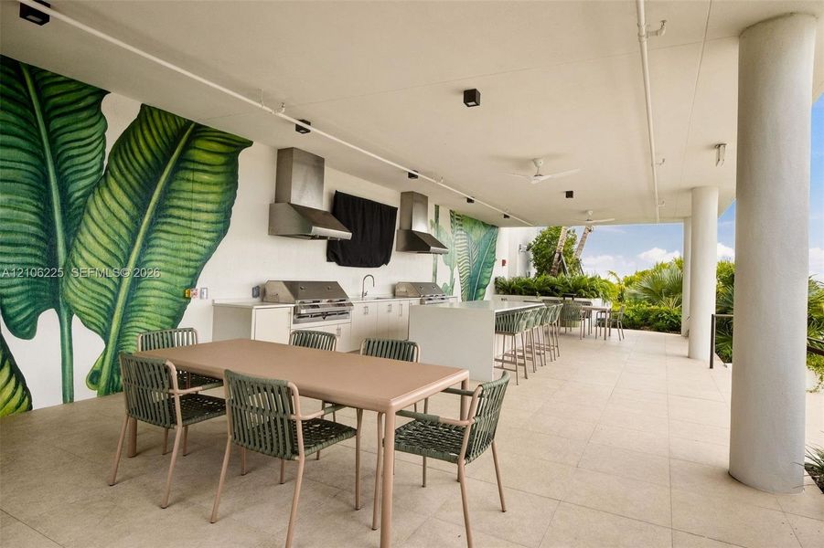 Covered outdoor kitchen featuring a stone-topped island, stainless steel built-in grills, overhead stainless steel vent hoods, large-format tile flooring, and integrated ceiling lighting Covered outdoor kitchen featuring a stone-topped island, stainless steel built-in grills, overhead stainless steel vent hoods, large-format tile flooring, and integrated ceiling lighting