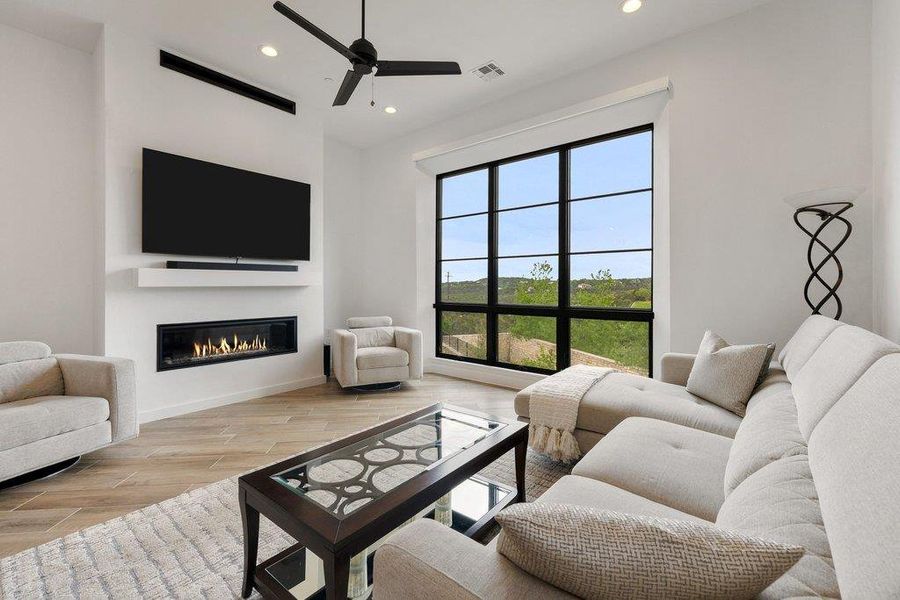 Open-concept living space with large black-grid windows, featuring a linear gas fireplace, built-in media shelf, and wood-finish flooring
