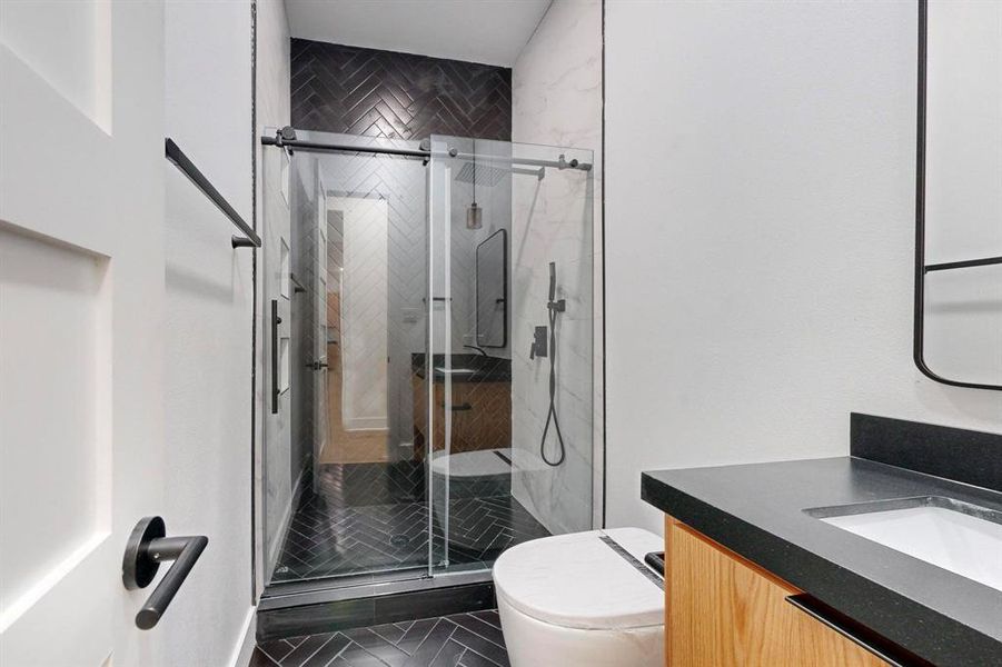 Contemporary bathroom featuring a glass-enclosed shower with black herringbone tile, a floating wood-finish vanity with a built-in sink and dark countertop, and matte black fixtures Contemporary bathroom featuring a glass-enclosed shower with black herringbone tile, a floating wood-finish vanity with a built-in sink and dark countertop, and matte black fixtures