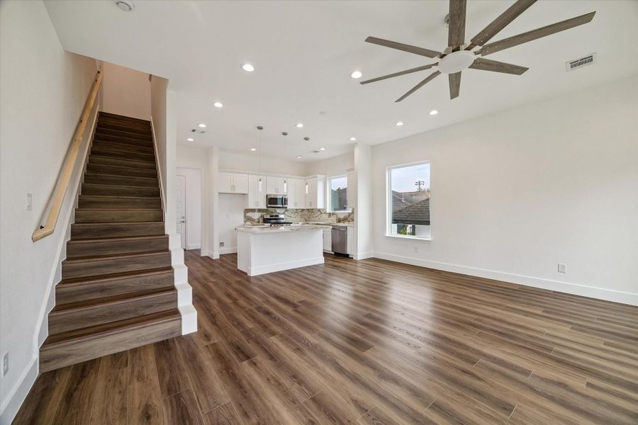 An expansive open-concept main level blends the living, dining, and kitchen areas into one inviting space. Rich wood-look flooring runs throughout, complemented by crisp white walls, recessed lighting, and a statement ceiling fan. The kitchen island anchors the room, while abundant windows bring in natural light and create a bright, contemporary setting. The flowing layout makes it easy to move between cooking, dining, and relaxing, creating a functional space for everyday life and entertaining. An expansive open-concept main level blends the living, dining, and kitchen areas into one inviting space. Rich wood-look flooring runs throughout, complemented by crisp white walls, recessed lighting, and a statement ceiling fan. The kitchen island anchors the room, while abundant windows bring in natural light and create a bright, contemporary setting. The flowing layout makes it easy to move between cooking, dining, and relaxing, creating a functional space for everyday life and entertaining.