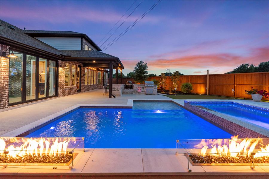 Another angle of the modern swimming pool, fire features and an outdoor kitchen. The space is beautifully lit, perfect for evening gatherings.