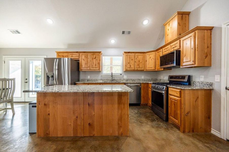 Kitchen with stainless steel appliances, finished concrete flooring, light stone counters, a kitchen island, and vaulted ceiling