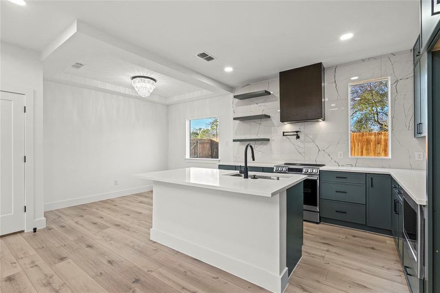 Kitchen with decorative backsplash, open shelves, stainless steel electric stove, an island with sink, and light stone countertops
