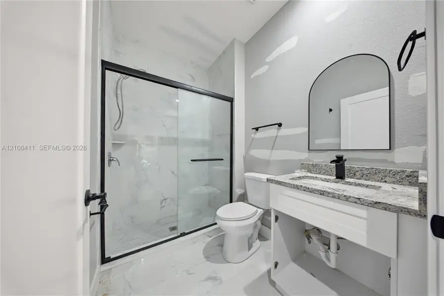 Bathroom featuring a walk-in shower with glass enclosure, stone-finish vanity countertop, arched wall mirror, and matte black hardware