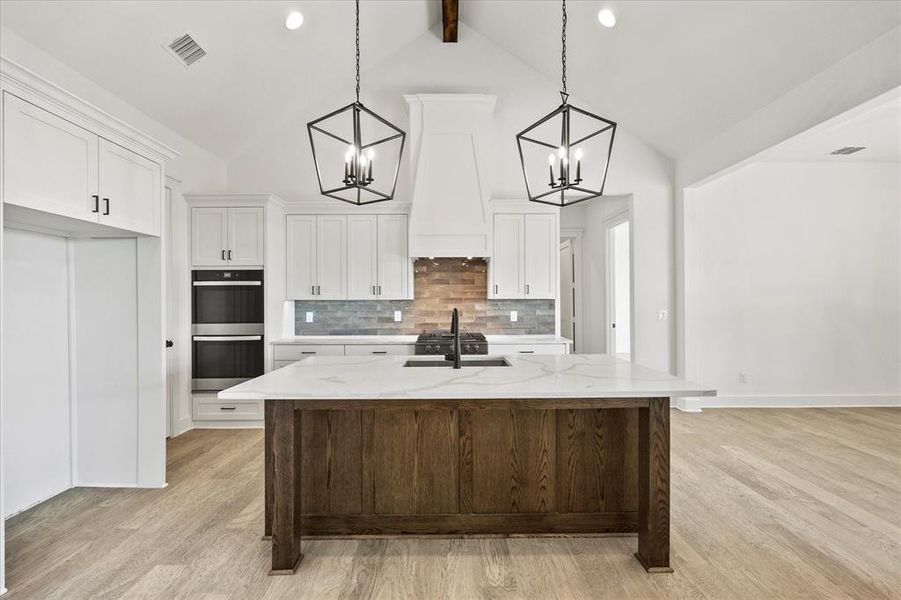 Kitchen featuring double oven, light stone countertops, a center island with sink, and lofted ceiling with beams