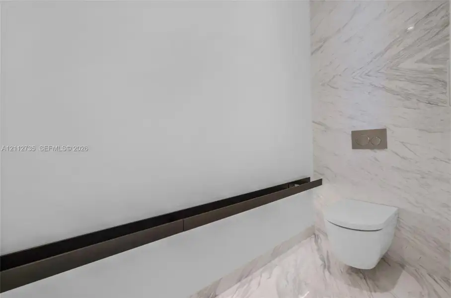Wall-mounted toilet with a dual-flush actuator, floor-to-ceiling stone-patterned tile wall, and a horizontal dark metal accent trim