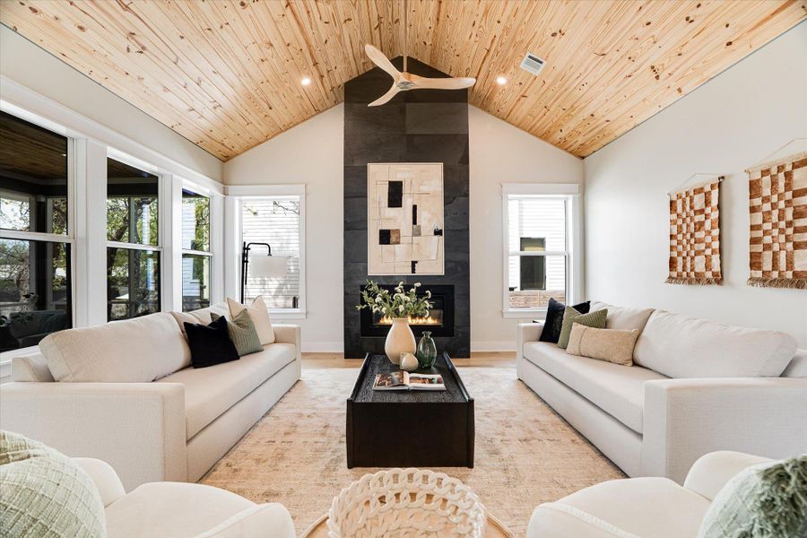 Living area featuring a vaulted wood-paneled ceiling, a modern fireplace with a dark stone facade, and large windows providing natural light Living area featuring a vaulted wood-paneled ceiling, a modern fireplace with a dark stone facade, and large windows providing natural light