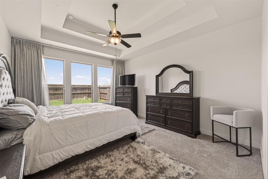 This downstairs owners' retreat features a cozy atmosphere with neutral tones, a plush carpet, and ample natural light from large windows. It includes a ceiling fan, elegant furniture with a dark finish, and a view of the backyard.