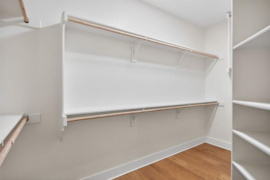 Walk-in closet featuring wood-finish flooring, white built-in shelving, white walls, and white baseboards Walk-in closet featuring wood-finish flooring, white built-in shelving, white walls, and white baseboards
