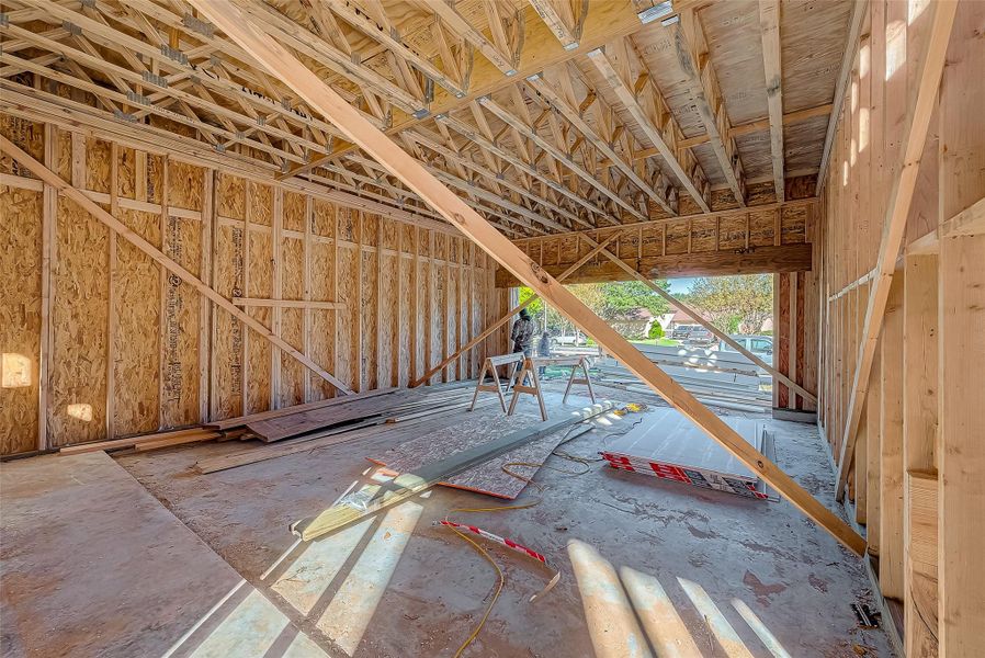 This photo shows a partially constructed room with exposed wooden framing and trusses. The open space suggests it might be a garage or large living area. Construction materials are scattered on the floor, and there's an opening leading outside, allowing natural light to enter.
